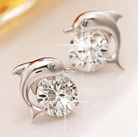 A pair of silver dolphin stud earrings, each featuring a large round clear gemstone and a small accent stone, displayed on a light cream surface with a soft-focus warm background.