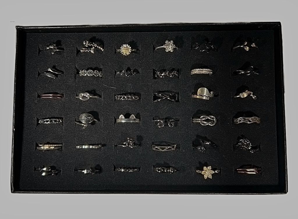A collection of silver and gold rings displayed in a black ring box with a black foam insert, photographed against a neutral gray background.