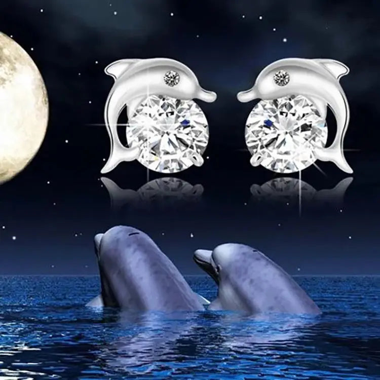 A pair of silver dolphin stud earrings with large clear round gemstones, shown against a dark ocean-and-moon background with dolphins swimming below.