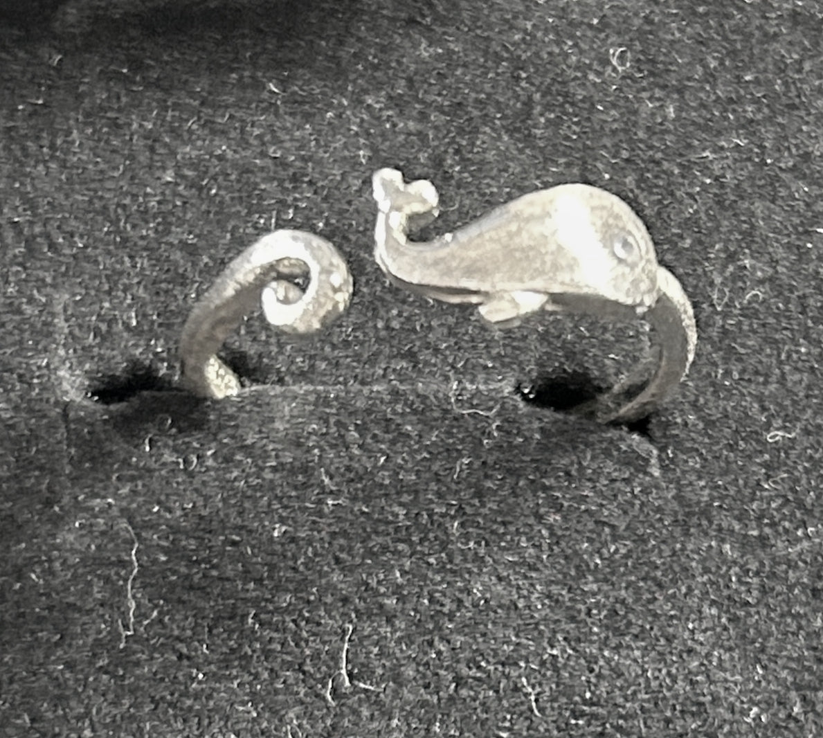 A pair of silver whale-shaped rings displayed on a dark textured velvet ring box.