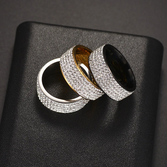 Three wide band rings with pavé-set clear stones, shown overlapping on a black textured surface. The rings are silver-toned, gold-toned, and black-toned.