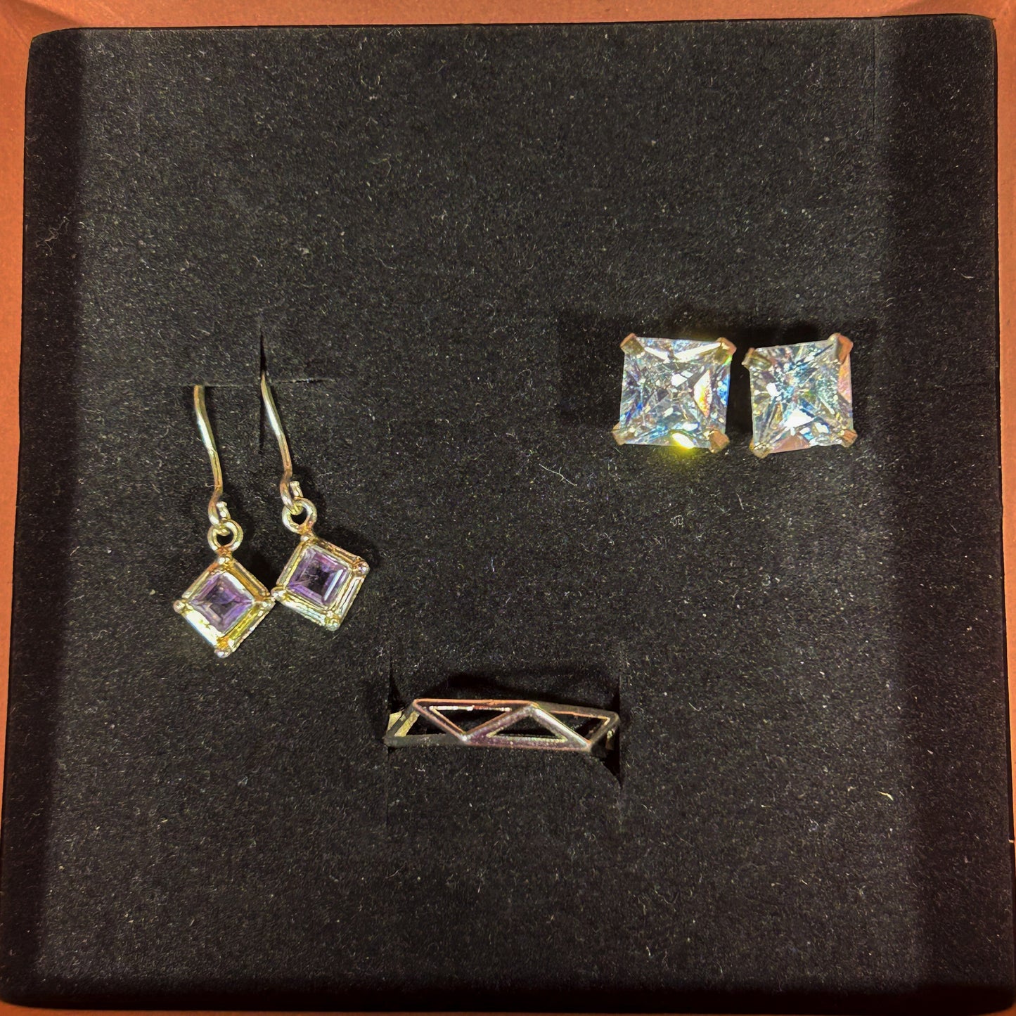 A jewelry set displayed on a black velvet insert inside an open box with a peach-colored border. The set includes a pair of gold-tone square stud earrings with iridescent clear stones, a matching pair of gold-tone diamond-cut drop earrings with light purple stones, and a small gold-tone geometric bar pin.