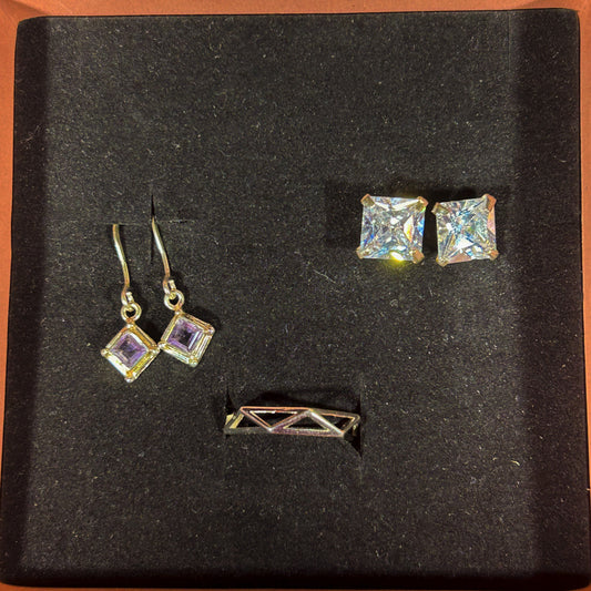 A jewelry set displayed on a black velvet insert inside an open box with a peach-colored border. The set includes a pair of gold-tone square stud earrings with iridescent clear stones, a matching pair of gold-tone diamond-cut drop earrings with light purple stones, and a small gold-tone geometric bar pin.