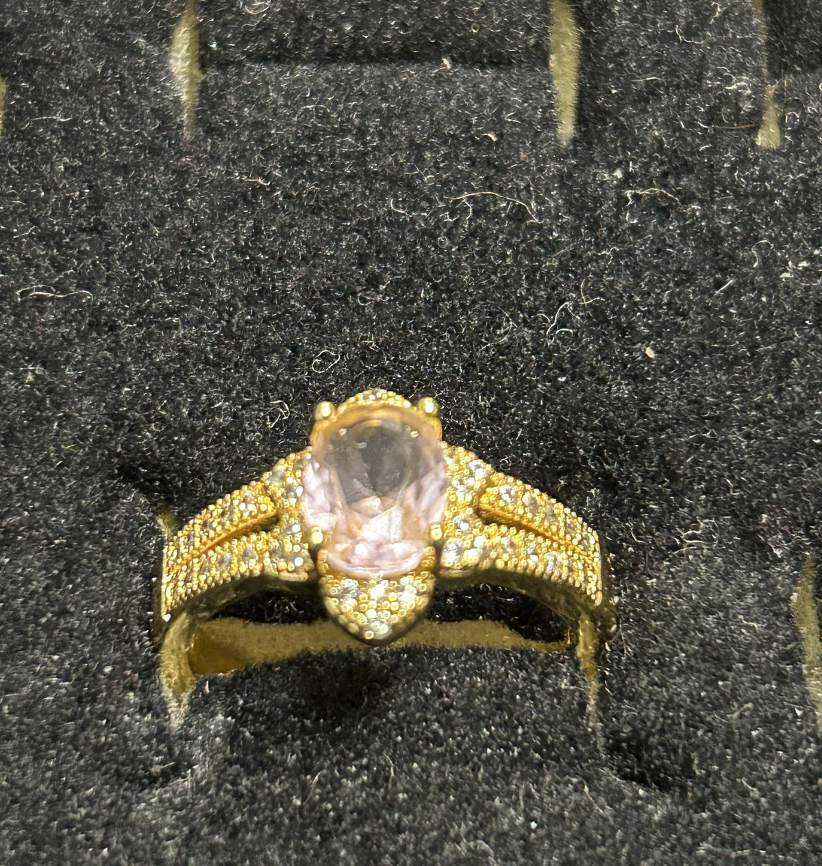 A gold ring with a large oval pink gemstone and pavé-set accent stones, displayed in a black textured ring box.