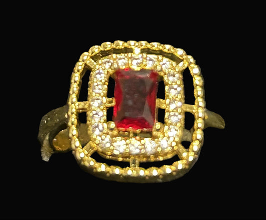 A gold ring with a square-shaped red gemstone surrounded by a double halo of small clear stones, displayed on a textured black surface.