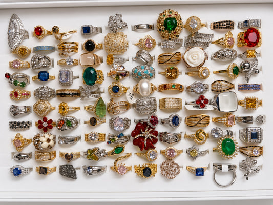 A large collection of assorted rings arranged in rows on a white background. The rings feature a wide variety of settings, metals (gold, silver, white gold), and colorful gemstones including green, red, blue, pink, and clear stones.