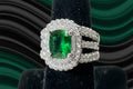 Emerald ring with diamond accents on a black stand against a green and black gradient background