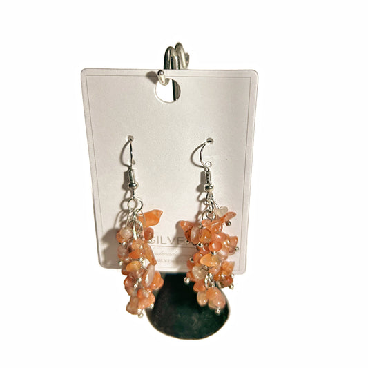 A pair of dangling earrings with clustered peach-orange stone beads and a dark green oval stone, attached to silver hooks and displayed on a white card.