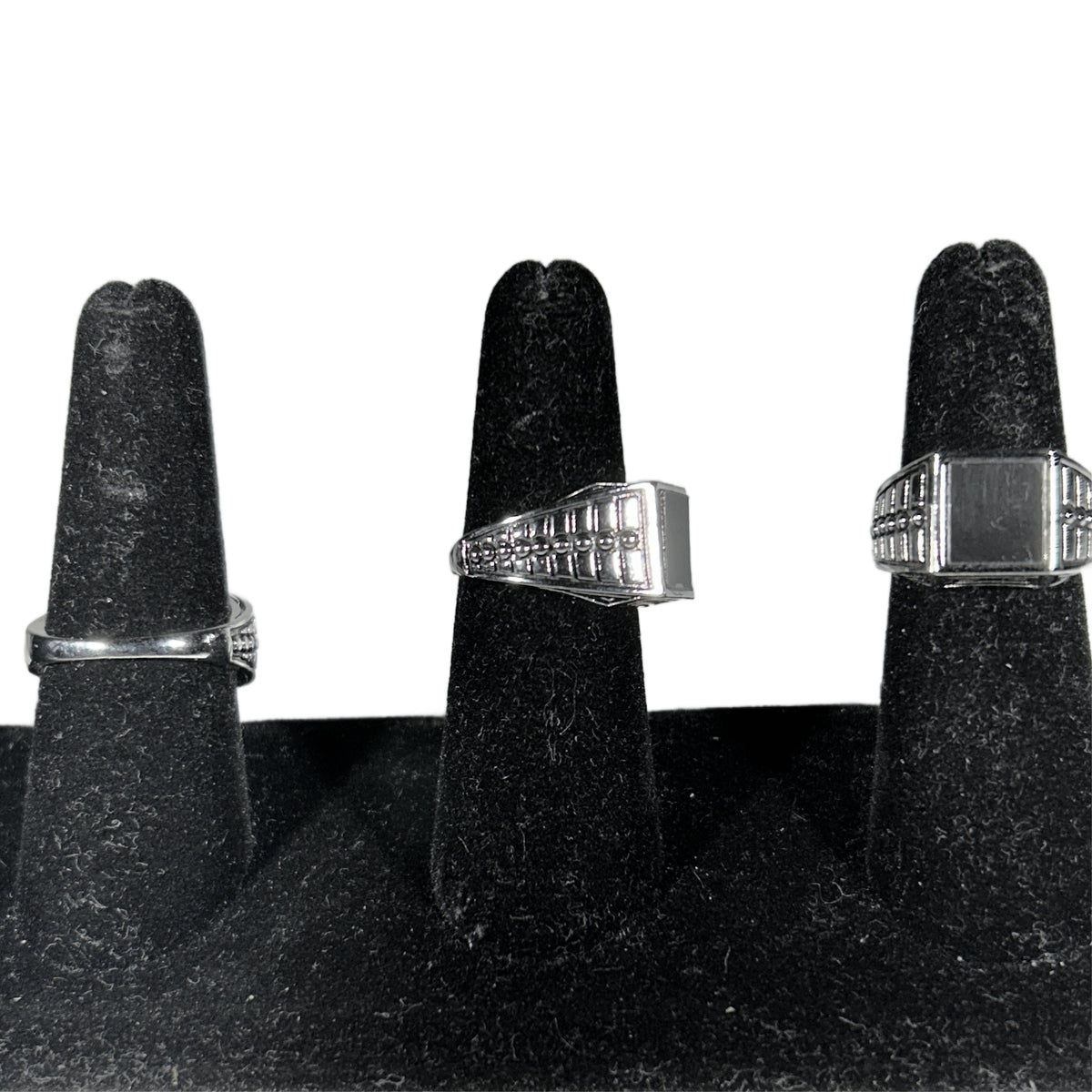 Three silver-toned men's rings with geometric and block designs displayed on black ring stands against a white background.