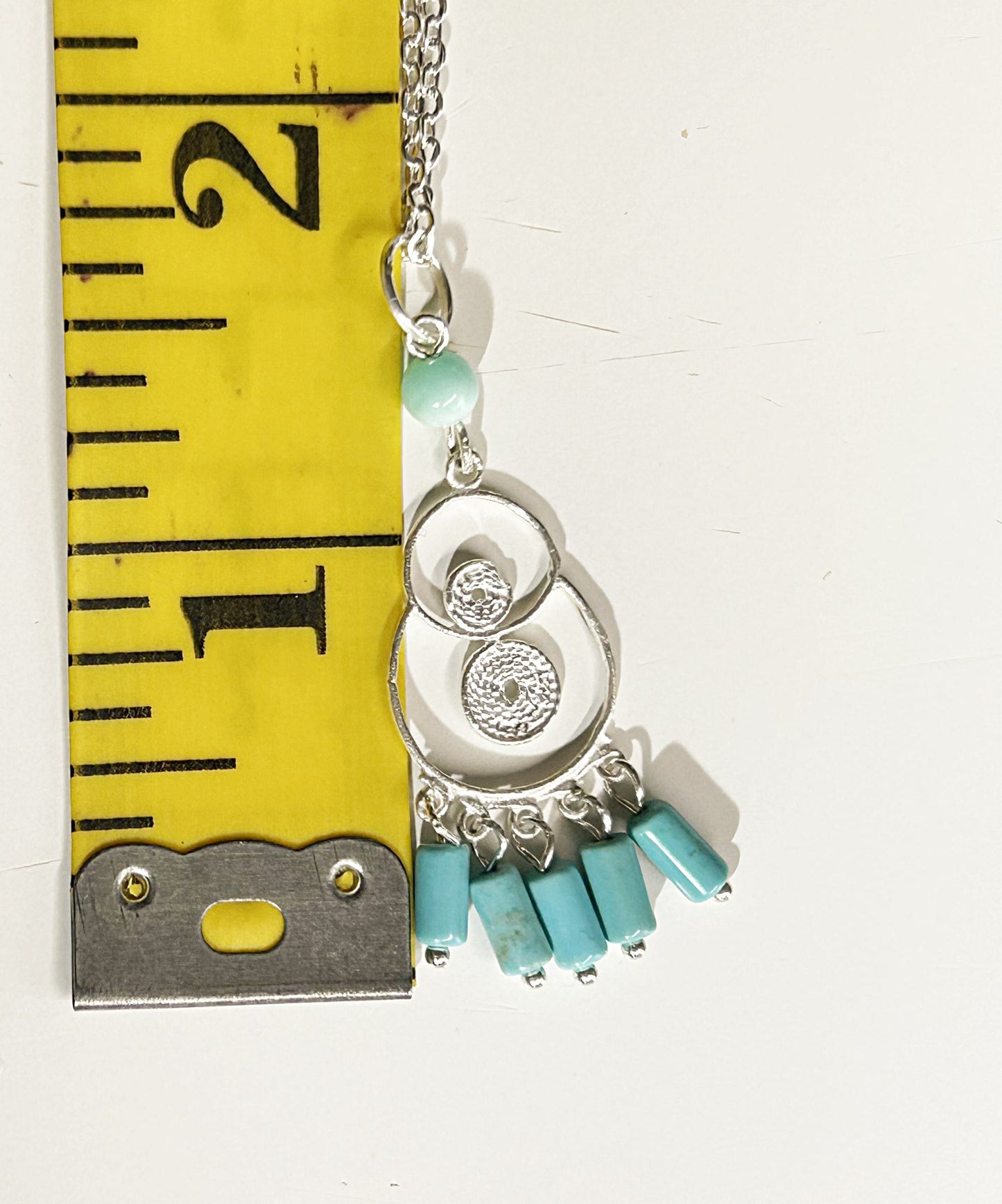 Necklace with turquoise beads and silver charms next to a measuring tape on a white background